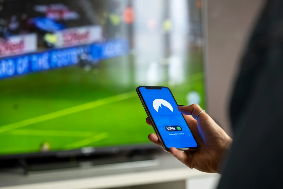 Hand holding smartphone with streaming app library in front of TV — illustrating multi-device IPTV access